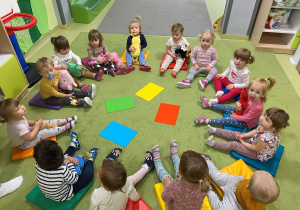 Dzieci siedzą w kole na dywanie i oglądają edukacyjne plansze z kolorami.