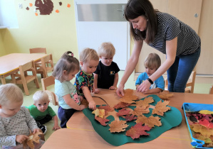 Dzieci z opiekunką przyklejają liście do drzewa na brystolu.
