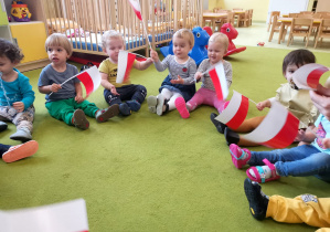Dzieci siedzą na dywanie trzymają chorągiewki biało-czerwone w rękach.