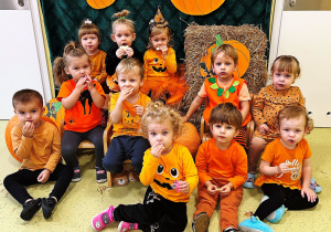 Dzieci są przebrane na pomarańczowo. Część z nich siedzi na krzesełkach a część na podłodze. Zdjęcie grupowe.