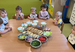 Dzieci siedzą przy drewnianym stoliku na którym stoją miseczki z pomidorem, sałatą, rzodkiewką i ogórkiem oraz taca z chlebem posmarowanym masełkiem. Przygotowują sobie śniadanie.