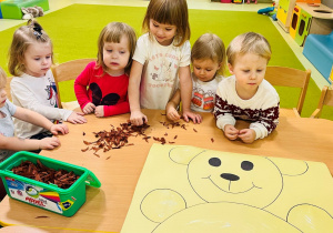 Dzieci stoją przy stoliku na którym leży brystol. Na nim narysowany jest pluszowy miś. Będą wyklejać go bibułą w kolorze brązowym.
