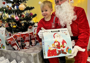 Olek siedzi na kolanie Mikołaja a w ręku trzyma prezent. Za Nimi są światełka a obok stoi kolorowo ustrojona choinka.