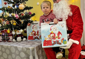 Franek siedzi na kolanie Mikołaja a w ręku trzyma prezent. Za Nimi są światełka a obok stoi kolorowo ustrojona choinka.