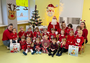 Zdjęcie grupowe. Wszyscy ubrani na czerwono siedzą z Mikołajem przy ubranej w kolorowe bombki choince.