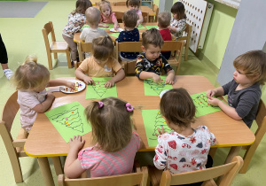 Dzieci siedzą przy stole i ozdabiają papierowe choinki kuleczkami z plasteliny.