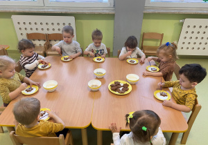 Dzieci siedzą przy stole i dekorują pierniczki lukrem i kolorowymi posypkami.