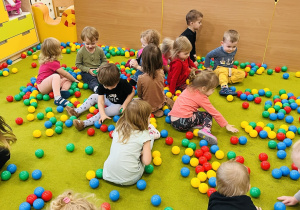 Dzieci siedzą na zielonym dywanie na którym są rozrzucone kolorowe kulki.