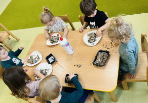 Dzieci siedzą przy drewnianym stoliku i przygotowują mini karminiki dla ptaków. Obtaczają papierowe rolki w ziarenkach.