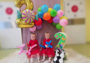 Ala i Hania siedzą na krzesełkach, obok stoi urodzinowa dwójka, wiszą dekoracje z balonów.
