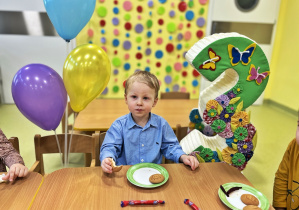 Franio siedzi przy stole obok niego stoi styropianowa trójka a z drugiej strony balony. Urodzinowy poczęstunek.