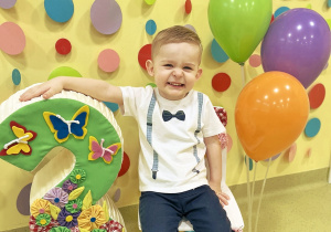 Olivierek siedzi na urodzinowym krześle obok niego stoją balony i urodzinowa trójka.