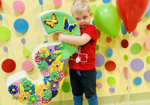 Oskarek stoi i trzyma styropianową trójkę oburącz. Obok niego stoją balony.