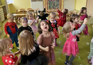 Dzieci w przebrane za różne postacie z bajek tańczą na dywanie a wśród nich lecą bańki