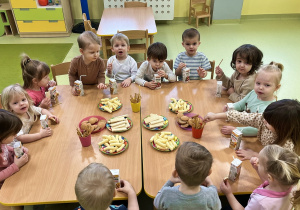 Dzieci siedzą przy połączonych drewnianych stolikach i zajadają urodzinowe przekąski.