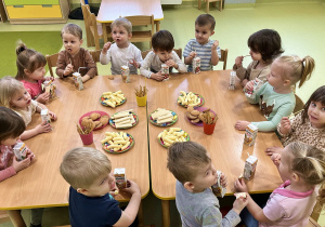 Dzieci siedzą przy połączonych drewnianych stolikach i zajadają urodzinowe przekąski.