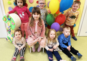 Dzieci stoją i siedzą obok Laury. Razem z nimi stoi trójka i balony. Zdjęcie grupowe.