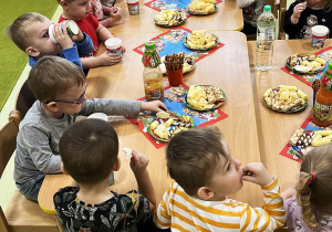 Dzieci siedzą przy połączonych drewnianych stołach i zajadają urodzinowe smakołyki.