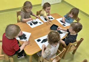 Dzieci malują kosmos na białej kartce granatową farbą. Siedzą przy drewnianym stoliku.