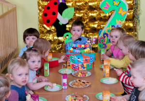 Dzieci siedzą przy stolikach częstując się urodzinowym poczęstunkiem.