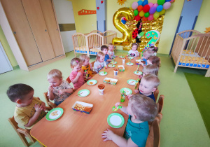 Ala z dziećmi siedzi przy stolikach podczas urodzinowego poczęstunku.
