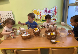 Dzieci siedzą przy stole i ozdabiają styropianowe jajka posypką z soli i kredy.