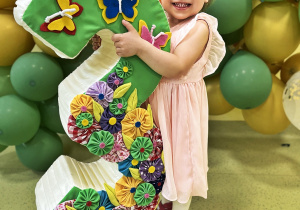 Julianka stoi z urodzinową trójką ozdobioną motylkami i kwiatkami za nią są zielono złote balony.