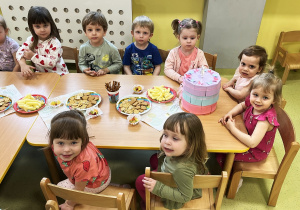 Dzieci siedzą przy drewnianym stoliku na którym leżą urodzinowe słodkości.