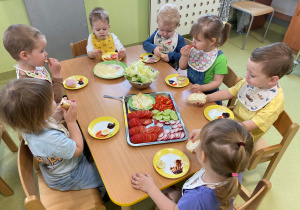 Dzieci siedzą przy stole i wykonują kolorowe kanapki.