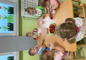 Dzieci siedzą przy stole piją koktajl truskawkowy i jedzą soczyste owoce.