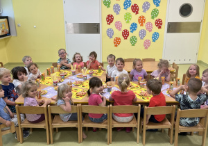 Dzieci siedzą przy dużym, wspólnym, drewnianym stoliku zajadając smakołyki z okazji dnia dziecka.