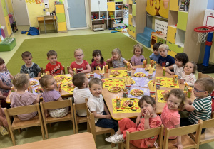 Dzieci siedzą przy dużym, wspólnym, drewnianym stoliku zajadając smakołyki z okazji dnia dziecka.