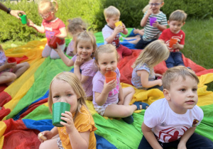 Dzieci siedzą na chuście animacyjnej na dworze i piją urodzinowy soczek.