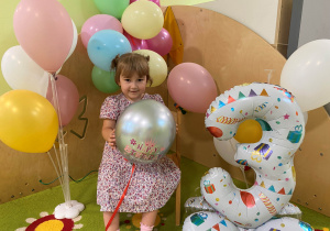 Helenka siedzi na krześle i pozuje do zdjęcia z kolorowymi balonami.