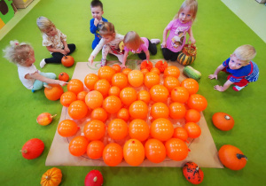 Dzieci przyklejają pomarańczowe baloniki na dużym szarym papierze na którym narysowany jest kształt dyni.