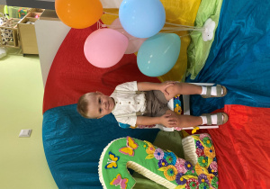 Filip siedzi na urodzinowym krzesełku a obok stoi cyfra 2. Z drugiej strony znajdują się kolorowe balony.