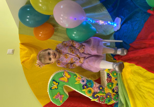 Ala siedzi na urodzinowym krześle obok jest cyfra 2. Z drugiej strony znajdują się kolorowe balony.
