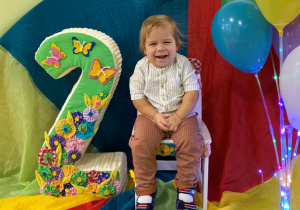 Kubuś siedzi na urodzinowym krześle a obok stoi styropianowa cyfra 2. Z drugiej strony znajdują się kolorowe balony.