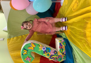 Hania siedzi na urodzinowym krześle. Obok stoi cyfra 2, ozdobiona motylkami i kwiatkami. Z drugiej strony znajdują się kolorowe balony.