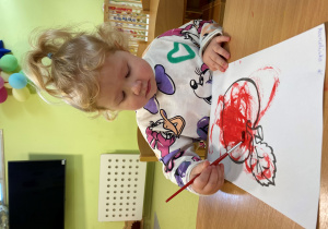 Marcelina maluje czerwoną farbą jabłko.