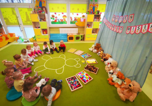 Dzieci siedzą na dywanie, na którym widnieje postać misia i różne materiały sensoryczne na tackach.