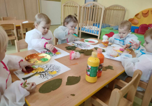 Dzieci siedzą przy stole i wykonują prace plastyczną przy użyciu farb.