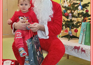 Szymon siedzi na kolanie Mikołaja. W ręku trzyma prezent. Za nimi stoi kolorowa choinka.