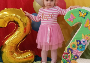 Ala stoi na urodzinowej chuście. Obok niej stoi styropianowa cyfra dwa oraz balon z cyfrą dwa.