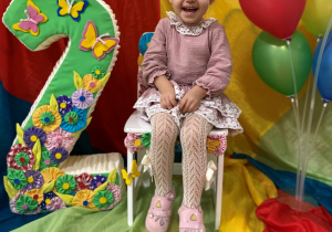 Nadia siedzi na urodzinowym krześle. Obok stoi styropianowa cyfra dwa oraz kolorowe balony.