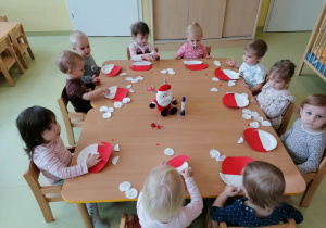 Dzieci siedzą przy stołach i naklejają płatki kosmetyczne na brodę Mikołaja.