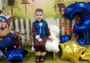 Eldar siedzi na urodzinowym krześle. Obok stoi cyfra trzy oraz leżą kolorowe balony.