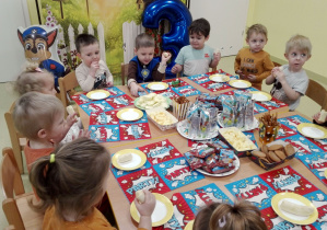Dzieci siedzą przy stoliku i jedzą urodzinowe smakołyki.
