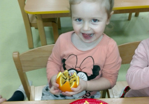Ala siedzi przy stoliku. W ręku trzyma pomarańcze z goździkami.