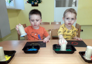 Tymek i Franek siedzą przy stoliku. W ręku trzymają rolkę papieru, którą maczają w farbie.